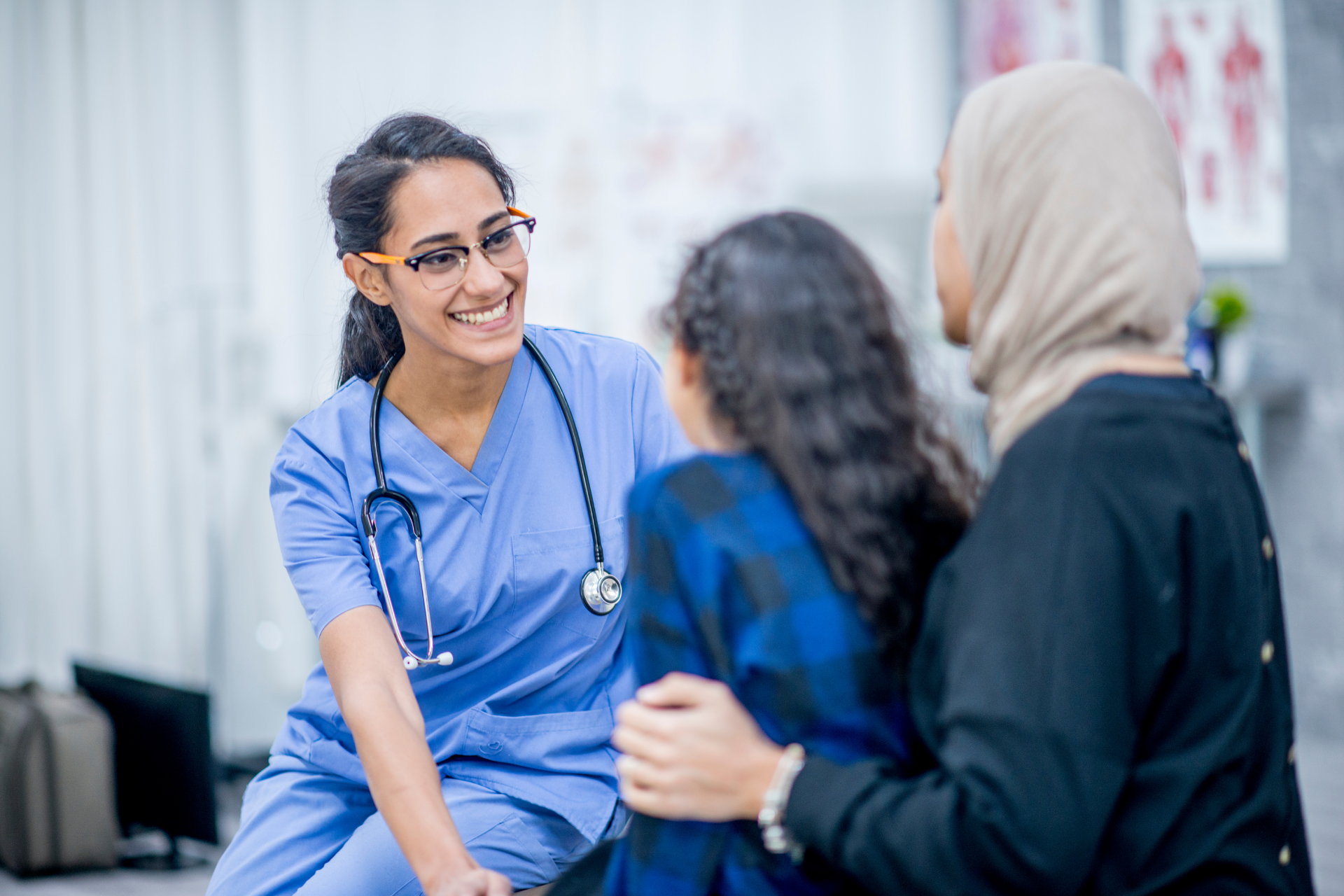 A woman in a head scarf and her young daughter sit and speak with a smiling female doctor with blue scrubs and a stethescope.