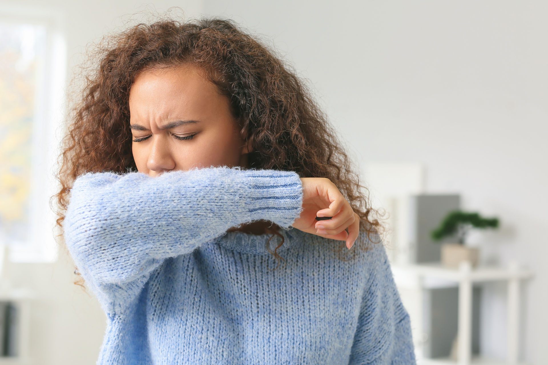 A woman with curly hair and a blue sweater coughs into the crook of her elbow.